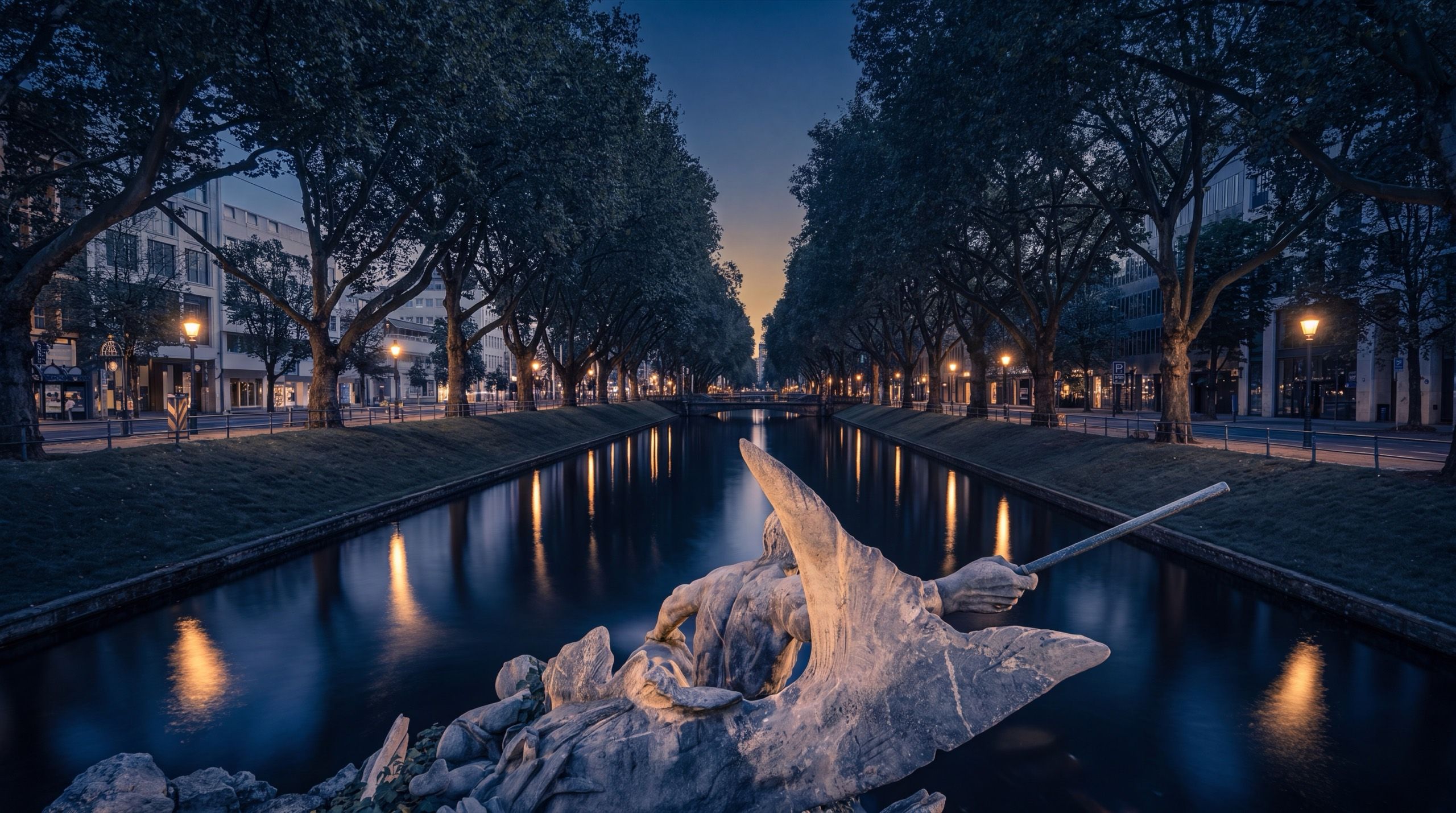 Düsseldorf's Königsallee at dusk: a tree-lined canal reflecting golden lamplight, the Triton fountain in the foreground, and the boulevard receding to a sunset glow.