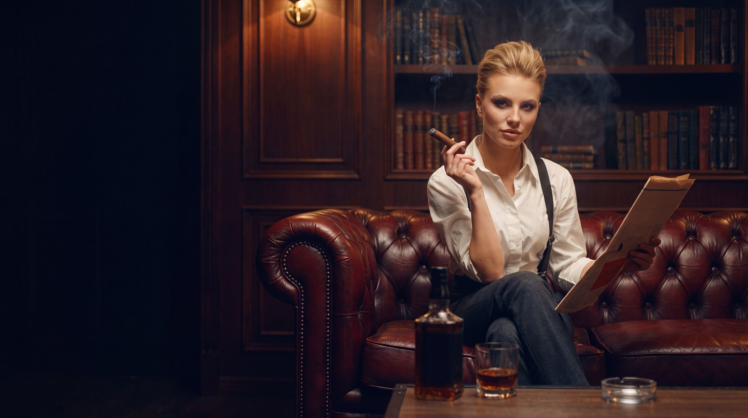 A woman on a leather chesterfield in a members' club library, reading a newspaper with a cigar in hand, whisky on the table and walls of books behind her.