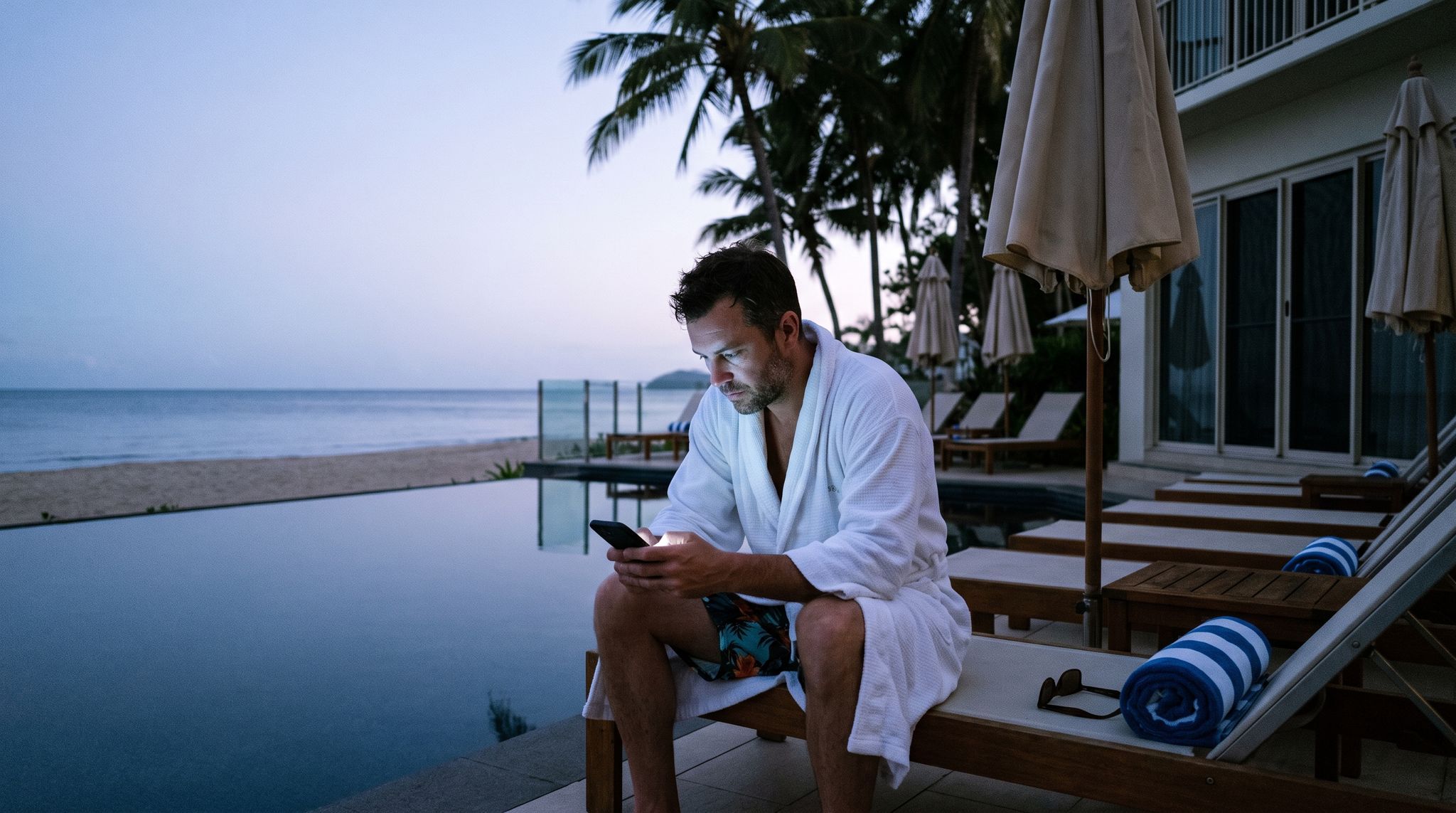 A man in a resort bathrobe types on his phone at dawn beside a still pool while his family sleeps inside.